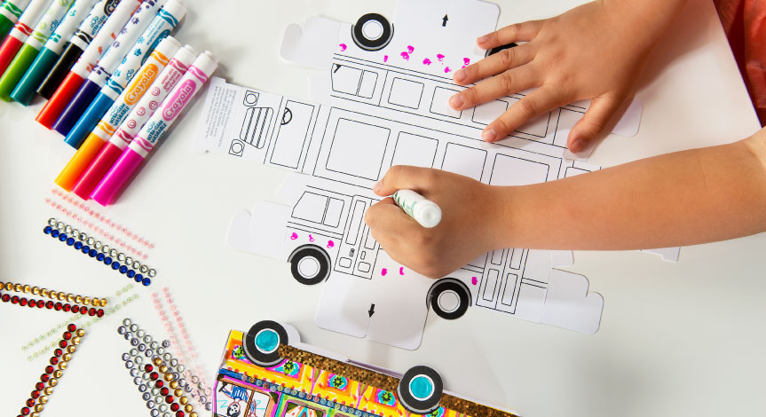 Child coloring a paper school bus on the table, using multiple different colored crayola markers.