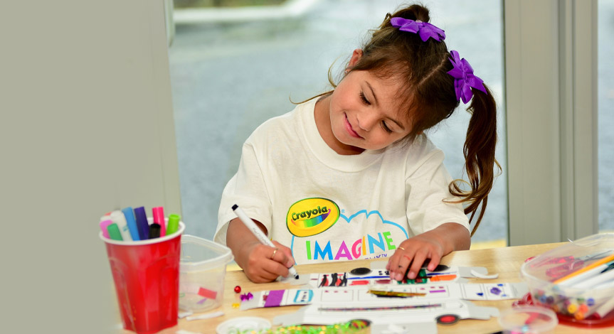 Girl wearing 2 purple hair bows and a Crayola Imagine Arts Academy Tshirt coloring during an art class